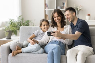 Mutlu genç ebeveynler ve tatlı kız çocuk akıllı telefondan içeriği izliyorlar, internetten iletişim için alet kullanıyorlar, aile selfie 'sini evdeki kanepede çekiyorlar, internet teknolojisinin tadını çıkarıyorlar.