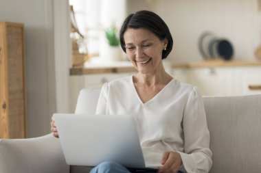 Neşeli, oldukça yaşlı, emekli iş kadını evinde dizüstü bilgisayar kullanıyor, rahat bir koltukta dinleniyor, video görüşmeleri yapıyor, ekrana bakıyor, gülümsüyor, gülüyor, çevrimiçi iletişimden keyif alıyor.