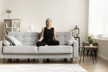 Ciddi, sakin, orta yaşlı bir kadın evde meditasyon yapıyor, gözleri kapalı koltukta oturuyor, lotus pozunu koruyor, zen eller, konsantrasyon uyguluyor, stres atağı yapıyor, rahatlık için gevşiyor.