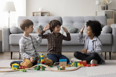 Diverse team of cheerful kids playing game on home floor, constructing towers, city road from toy building bricks, giving group high five, celebrating teamwork success clipart