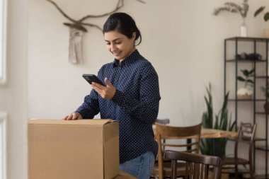 Evde karton kutu paketleyen genç Hintli kadın, cep telefonunu kullanıyor, kurye, teslimat servisi arıyor, internet mağazasından paketleri açıyor.