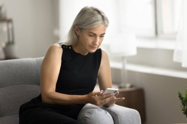 Akıllı telefonlu ciddi bir son sınıf öğrencisi kanepede oturuyor, cep telefonuyla yazı yazıyor, mesaj yazıyor, cep telefonu kullanıyor, iletişim için internet uygulaması kullanıyor, internetten alışveriş yapıyor.