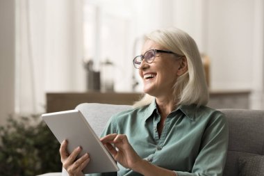 Neşeli, yaşlı bir kadın tabletle kanepede dinleniyor, emekliliğin tadını çıkarıyor, modern teknoloji kullanıyor, internet iletişimi kuruyor, dijital cihazı tutuyor, başka yere bakıyor, gülümsüyor, gülüyor.