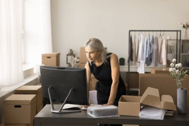 Küçük depolama yerinde masaüstü bilgisayarını kullanan, notlar yazan, işlem sıralayan, web mağazasından alışveriş yapan, paketleme, paketleme ve paketleme için ürün hazırlayan olgun bir internet mağazası sahibi.