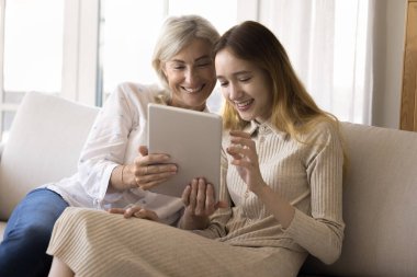 Neşeli ergen torun kız ve elinde tablet olan mutlu büyükanne evde kablosuz internet teknolojisi kullanıyor iletişimin tadını çıkarıyor, gülümsüyor, içeriği izliyor, online alışveriş yapıyor.