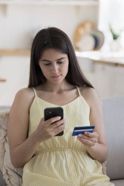 Banka müşterisi kız cep telefonu ve kredi kartıyla online ödeme yapıyor, ev koltuğunda oturuyor, cep telefonuyla yazı yazıyor, internet üzerinden alışveriş için ticari finansal uygulamayı kullanıyor.