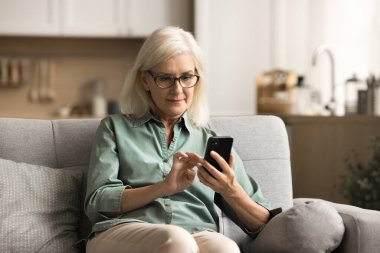 Evde cep telefonuyla yazılar yazan, sağlık hizmetlerini akıllı telefondan kullanan, kablosuz internet bağlantısı olan, evdeki kanepede oturan ciddi, yaşlı ve göz giysisi giyen bir kadın.