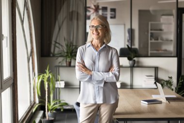 Neşeli, düşünceli, profesyonel, iş fikirleri üzerine düşünen şirket geleceği vizyonu olan, elleri çapraz bir şekilde ofiste duran, pencereye bakan, gülümseyen bir kadın. CEO, iş sahibi portresi