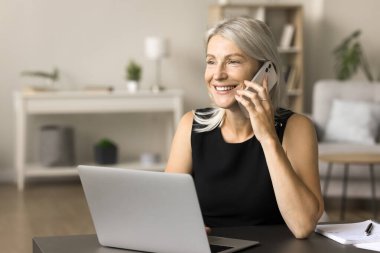 Mutlu orta yaşlı girişimci kadın cep telefonuyla dizüstü bilgisayarla konuşuyor, evden çalışmak için kablosuz internet bağlantı teknolojisini kullanıyor, gülümsüyor, cep telefonu konuşmalarından keyif alıyor.