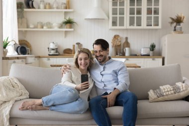 Mutlu milenyum çifti rahat bir evde rahat vakit geçiriyor, akıllı telefon kullanıyor, selfie çekiyor, video konferansında konuşuyor, gülümsüyor, gülüyor, kanepede dinleniyor.