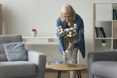 Çekici, olgun bir kadın, güzel bir buket çiçek hazırlıyor, rahat oturma odasındaki sehpanın üzerine vazo koyuyor, estetik cazibe ve doğanın dokunuşunu, güzelliği ve tazeliği modern yaşam alanına ekliyor.
