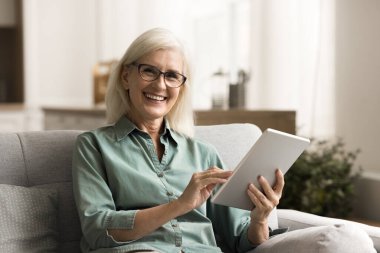 Gözlüklü, tabletli, kameraya bakan, gülümseyen, gülen, ev resmi için poz veren mutlu, son sınıf güzeli bir kadın. Emekli öğrenci internetteki eğitimden zevk alıyor, elinde aletle kanepede oturuyor.