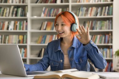 Kablosuz kulaklıklarla el sallayan neşeli, genç, kızıl saçlı kız laptopa bakıyor, gülümsüyor, açık kitaplarla üniversite kütüphanesinden video konuşmaları yapıyor. Kafasından vurulmuş.