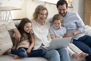 Heyecanlı ebeveynler ve iki mutlu çocuk evde internet iletişimi için dizüstü bilgisayar kullanıyorlar, kablosuz internet bağlantısı, kanepede birlikte dinleniyorlar, film izliyorlar, gülümsüyorlar, gülüyorlar.