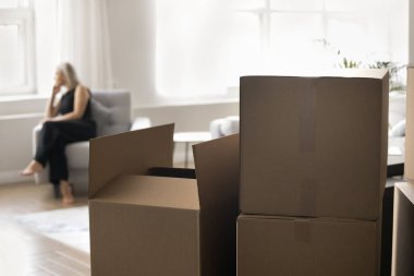 Nakliye, kargo, nakliye ve ulaşım için karton kutulara odaklanın. Siyah elbiseli üzgün kadın, bulanık arka planda, evin iç tarafındaki pencereye bakıyor.