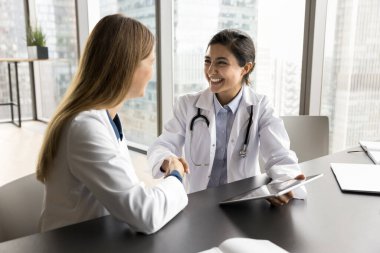 Neşeli, çeşitli klinik meslektaşları. Çalışma masasında el sıkışan, gülümseyen, gülen kadınlar. Genç pratisyen, mutlu tıp uzmanına şükranlarını sunarak el sıkışırken, yardımları için teşekkür ediyor.