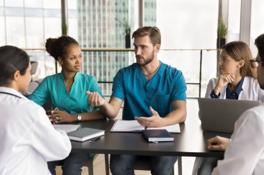 Mavi üniformalı ciddi yakışıklı doktor iş toplantısında çok ırklı iş arkadaşlarıyla konuşuyor, hastane çalışanlarının işini tartışıyor, uzmanlık yapıyor, pratisyenlere danışmanlık yapıyor.