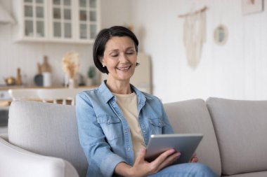 Mutlu, rahatlatılmış, medya içeriğini izleyen, tablet bilgisayarda film izleyen, evdeki koltukta oturan, ekrana bakan, gülümseyen, gülen, video konuşması yapan, konferanstan zevk alan yaşlı bayan.