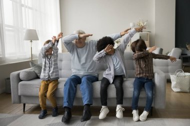 Aktif küçük torun çocukları ve yaşlı büyükbaba dans jesti yapıyor, koltukta oturuyor, poz veriyor, eğleniyor, yüzlerini dirseklerle kapatıyor. Büyükbaba ve çocuklar aile tatilinin tadını çıkarıyorlar.