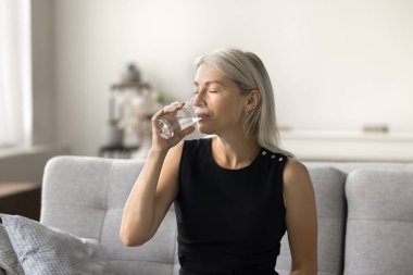 Barışçıl, güzel, olgun bir kadın sağlık sigortası için kapalı gözlerle doğal maden suyu içiyor. Sağlıklı metabolizma, ferahlatıcı, evdeki kanepede oturuyor, susuzluğunu gideriyor.
