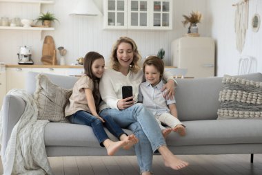 Olumlu blogcu anne evde iki küçük kardeş çocuğa sarılıyor, cep telefonunu internet iletişimi için kullanıyor, görüntülü konuşuyor, aile fotoğrafı çekiyor.