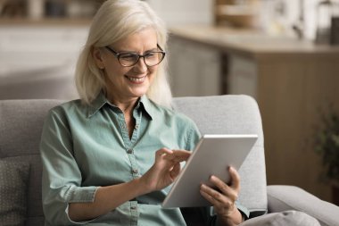 Gözlüklü yaşlı bir kadının dijital aletlerle evdeki koltukta internet iletişimi, uzaktan eğitim, internet içeriğini okumak, dokunmatik ekran, gülümseme, kahkaha