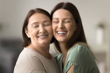 Mutlu kıdemli anne ve yetişkin kız yanaklarına dokunarak ve kapalı gözlerle sarılıyorlar. Olgun bir anneyi sevgi, minnettarlık, ilgi, gülümseme ve kahkahalarla kucaklayan olumlu bir genç kadın.