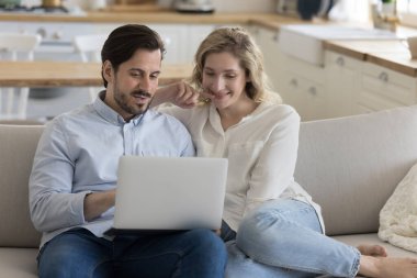 Kendine güveni tam yakışıklı bir adam, neşeli kız arkadaşına dizüstü bilgisayarında içerik gösteriyor, online başvuru işini açıklıyor. Milenyum çifti evlerinde bilgisayar kullanıyor, kanepeye yakın oturuyor.