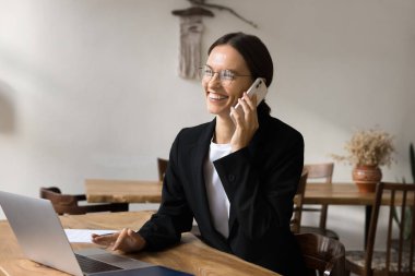 Resmi takım elbiseli neşeli genç iş kadını dizüstü bilgisayarda cep telefonuyla konuşuyor, dijital cihazlar kullanıyor, iş iletişimi için kablosuz teknoloji kullanıyor, iş sürecinden keyif alıyor, cep telefonuyla konuşuyor, gülüyor.