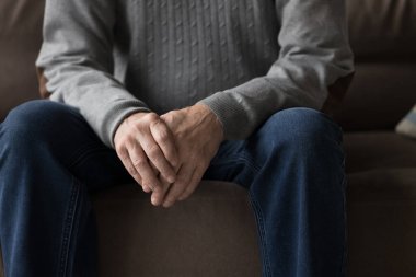 Son sınıf öğrencisi, rahat kıyafetlerle kanepede oturuyor, el ele tutuşuyor, avuç içlerini katlıyor, dirseklerini kalçalarına dayıyor, sohbet için poz veriyor, vücut hareketlerini bekliyor, endişe duyuyor. Erkek ellerine yakın dur.