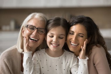 Mutlu küçük kız güzel büyükanne ve annenin yumuşak cildine dokunuyor. Neşeli büyükanne, anne ve küçük ergen çocuk birbirine yakın oturuyor dokunuşlar, kapalı gözler, dişlek gülümsemeler, kahkahalar