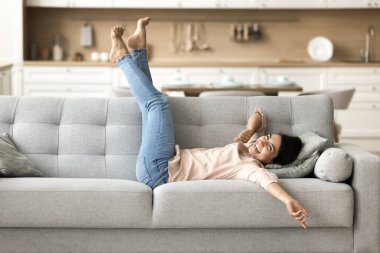Neşeli, güzel, genç Hintli bir kadın rahat koltukta uzanıyor, bacaklarını kaldırıyor, kameraya bakıyor, portre için gülümsüyor, gülüyor, eğleniyor, ev içinde keyif çatıyor.