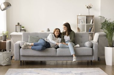 Olumlu anne ve ergen çocuk rahat bir koltukta dinleniyor, modern apartman dairesinde dinleniyor, rahat bir evde keyfine bakıyor, konuşuyor, sohbet ediyor, gülüyor. Tam uzunluk