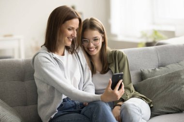 Neşeli anne ve genç kız birlikte akıllı telefondan selfie çekiyorlar, cep telefonuyla video görüşmesi yapıyorlar, gülümsüyorlar, gülüyorlar, sarılıyorlar, arkadaşlıktan keyif alıyorlar.