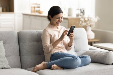 Neşeli mutlu Hintli kadın akıllı telefonunda medya uygulaması kullanıyor, ev koltuğunda eğleniyor, dijital cihazda daktilo çekiyor, kablosuz internet teknolojisinden hoşlanıyor, bağlantı kuruyor, sohbet ediyor.