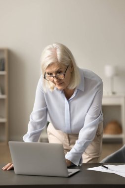 Evde çalışan, ofis masasında duran, dizüstü bilgisayarı kullanan, daktilo kullanan, uzak görevler için internet iletişimini kullanan, gözlüklü yaşlı, serbest çalışan yaşlı bir kadın.