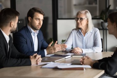 Resmi takım elbiseli kendine güvenen genç proje yöneticisi toplantı masasında meslektaşları ve yaşlılarla konuşuyor, çalışma planını, takım çalışması stratejisini, rapor verme, beyin fırtınası fikirlerini anlatıyor.