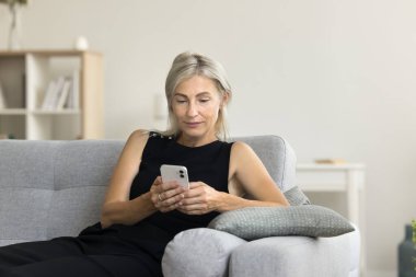 Sarışın, orta yaşlı bir kadın cep telefonuyla koltukta dinleniyor, akıllı telefondan mesaj atıyor, sohbet ediyor, daktilo ediyor, internet uygulaması kullanıyor.