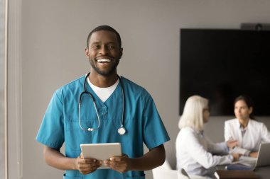 Neşeli yakışıklı Afrikalı Amerikalı doktor profesyonel portresi. Beyaz üniformalı genç siyah cerrah elinde dijital tablet cihazıyla takım odasında dikilip kameraya bakıyor.