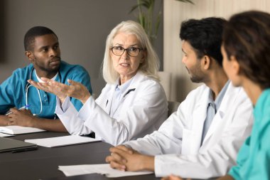 Ciddi kıdemli doktor toplantı masasında çeşitli hastane personeliyle konuşuyor, konuşuyor, talimatlar veriyor, tıbbi sorunları açıklıyor. Tıbbi uzman, genç stajyerleri eğiten akıl hocası.