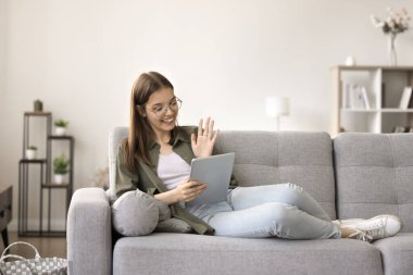 Pozitif zeki liseli kız video üzerinden konuşuyor tablet üzerinde sohbet ediyor, kanepe üzerinde dinleniyor selamlaşarak el sallıyor merhaba diyor, gülümsüyor, gülüyor, internet iletişiminden keyif alıyor.