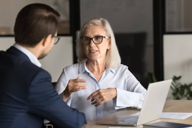 Ciddi, yaşlı, profesyonel bir kadın iş yerinde genç bir meslektaşla konuşuyor, birlikte proje üzerinde çalışıyor, işbirliği ve yönetim stratejisini tartışıyor. Ofis masasında iş ortakları konuşuyor.