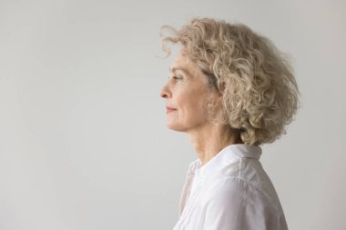 Düşünceli, ciddi, sarışın, olgun bir kadın beyaz stüdyo arka planında duruyor, derin düşüncelere dalıyor. Güzel, son sınıf model, kıvırcık, sarı saçlı bir portre.