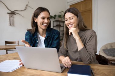 İki mutlu genç iş kadını dizüstü bilgisayardaki proje için yaratıcı stratejiyi tartışıyor, bilgisayarın başında oturuyor, gülümsüyor, gülüyor, online internet içeriğini izliyor, iş fikirleri üzerine beyin fırtınası yapıyor.