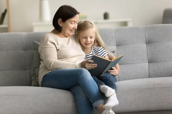 Positive caring grandma reading book to granddaughter kid on comfortable couch. Grandmother telling fairytale to little girl at home, teaching child to read, enjoying educational activity