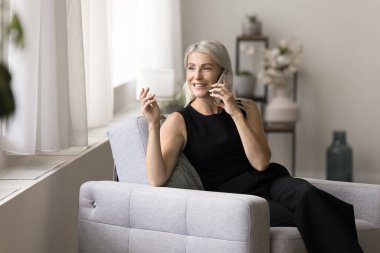 Mutlu, sarışın, olgun bir kadın cep telefonuyla konuşuyor, evdeki rahat koltukta dinleniyor, düşünceli bir yüz ifadesiyle başka yere bakıyor, cep telefonundan hoşlanıyor, konuşuyor, dinliyor, gülümsüyor.