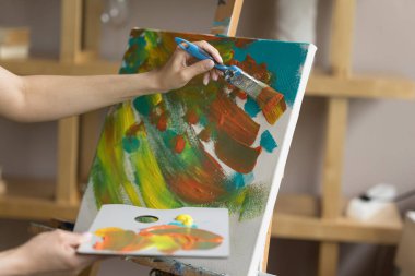Ressam kadının paleti akrilik boya ile kullanırken, boya fırçasıyla, tuval üzerinde fırça darbeleri yaparken. Stüdyoda çalışan sanat okulu öğrencisi, soyut resim çiziyor. Eller yukarı.