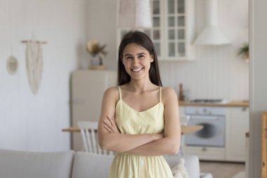 Kendinden emin, genç, esmer, kolsuz yazlık elbise giyen, kameraya çapraz ellerle bakan, gülümseyen, evde poz veren güzel bir kız. İç mekan kadın portresi