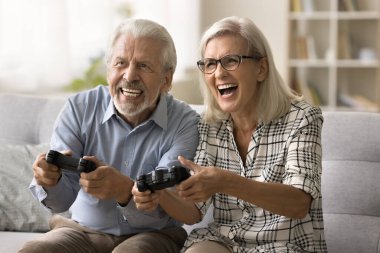 Neşeli, heyecanlı yaşlı çift çevrimiçi video oyunu oynuyor, konsoldaki joypad 'leri kullanıyor, kanepede oturuyor, bağırıyor, gülüyor, eğleniyor, aile eğlencesi, eğlenceli oyun zamanı