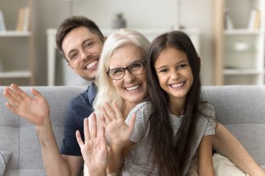 Neşeli küçük kız, mutlu büyükanne ve genç yakışıklı baba kameraya bakıyor, el sallıyor, birbirlerinin arkasında oturuyor, gülümsüyor, gülüyor, aile fotoğrafı için poz veriyor.